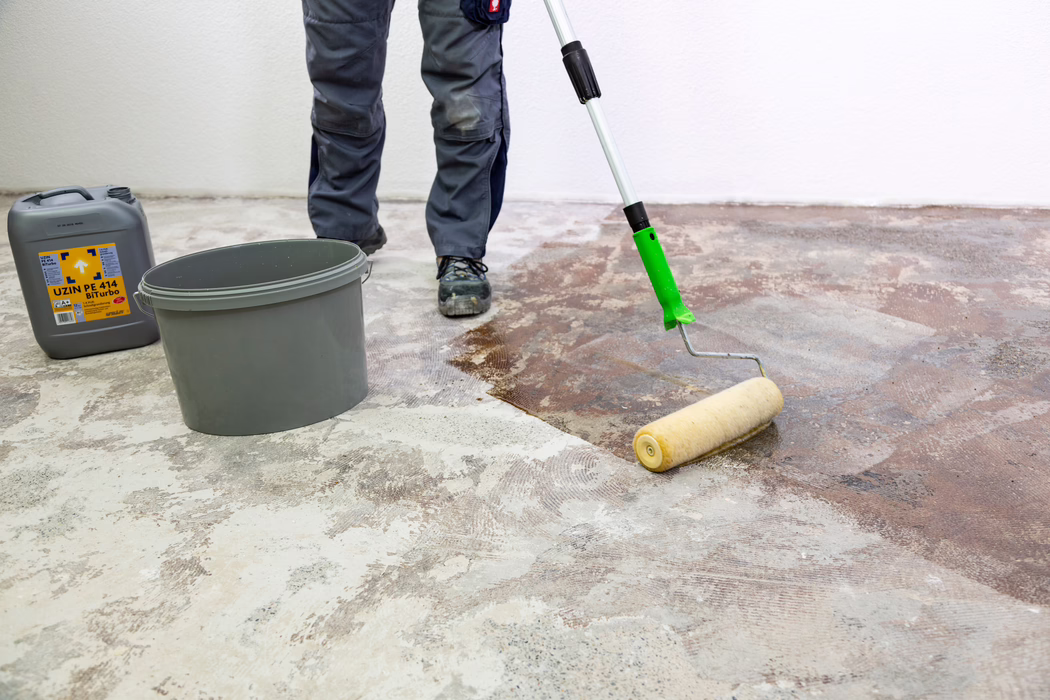 UZIN_Application Image_PE 414_9406_2018-11 En arbetares underben och skor syns när hen med en långskaftad färgrulle applicerar en brun primer eller lim på ett naket betonggolv. En grå plasthink och en grå dunk märkt "UZIN PE 414 Biturbo" står i närheten mot en vit vägg. Arbetaren bär grå arbetbyxor och svarta skyddsskor; golvet visar fläckar av gammalt lim och slipad betong medan rullen sprider vätskan jämnt för att förbereda underlaget.