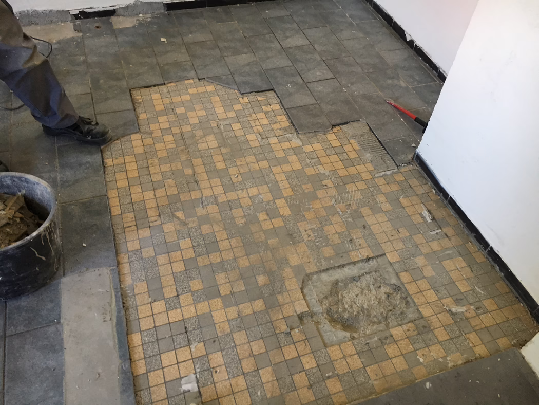 UZIN_Application_Image_UZIN NC 110_0445_ 2018-03 Delvis borttagen klinkergolv under renovering: i förgrunden syns små beige‑grå mosaikplattor med slitna och saknade partier; större mörka rektangulära plattor vid kanterna har lyfts och avslöjar lim och underlag. Till vänster syns en arbetares ben och svarta skor bredvid en hink med borttagna plattor; till höger lutar en röd kofot mot väggen. Vita målade väggar med svart sockel, damm och byggdamm spritt över golvet. Bild tagen från låg vinkel nedåt.