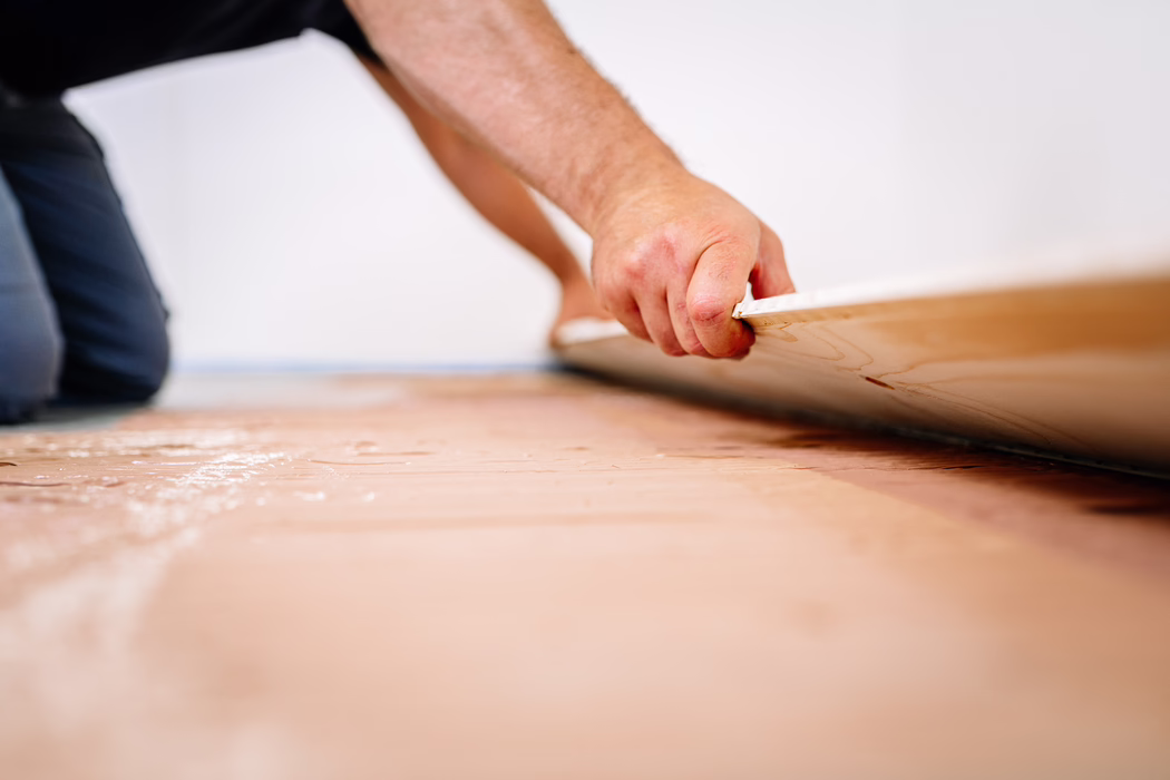 UZIN_Application_Image_MK_250_11_2021-11 Close-up of a person installing flooring: a kneeling worker's hand grips the edge of a laminate or wooden plank as it is being placed onto a prepared subfloor. The shallow depth of field emphasizes the textured plank edge and the hand, while the background shows a white wall and the worker's kneeling posture. The surface beneath appears to be an underlayment or adhesive layer with a slightly pink hue, and the image conveys careful manual work during a home renovation.